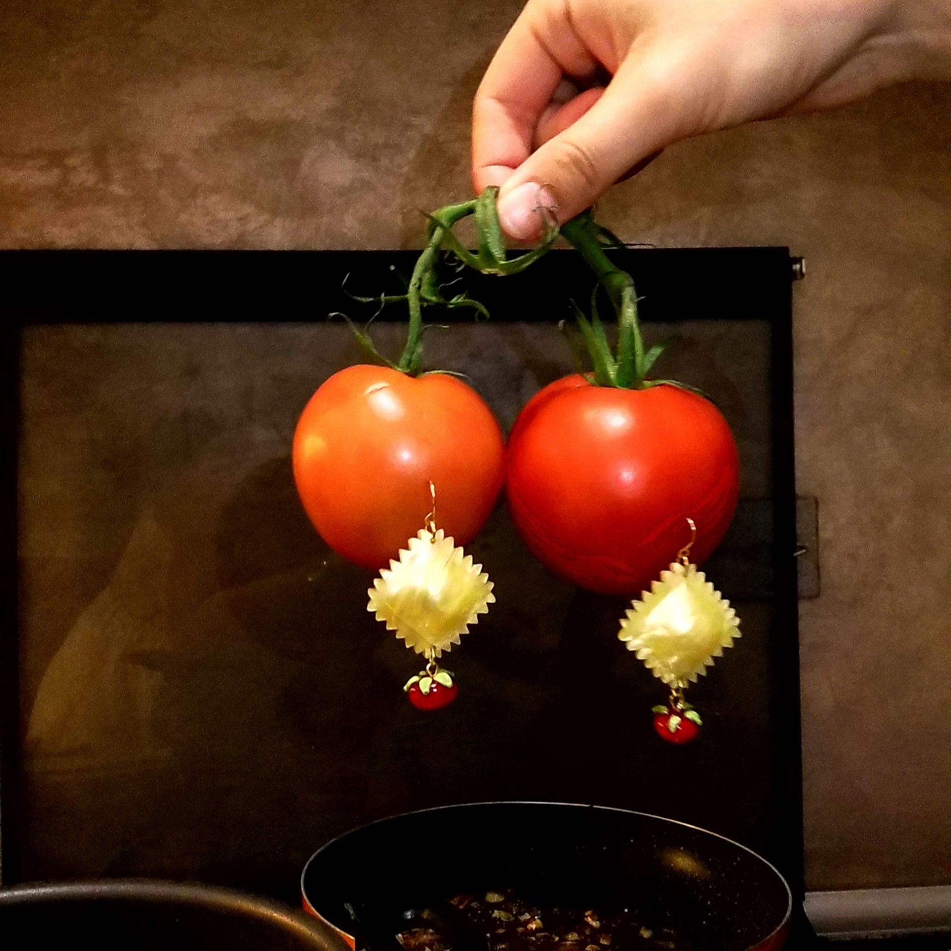 A pair of yellow pearlescent acrylic earrings designed to resemble tomatoes, with a person's hand holding them up to a pan on a stove.