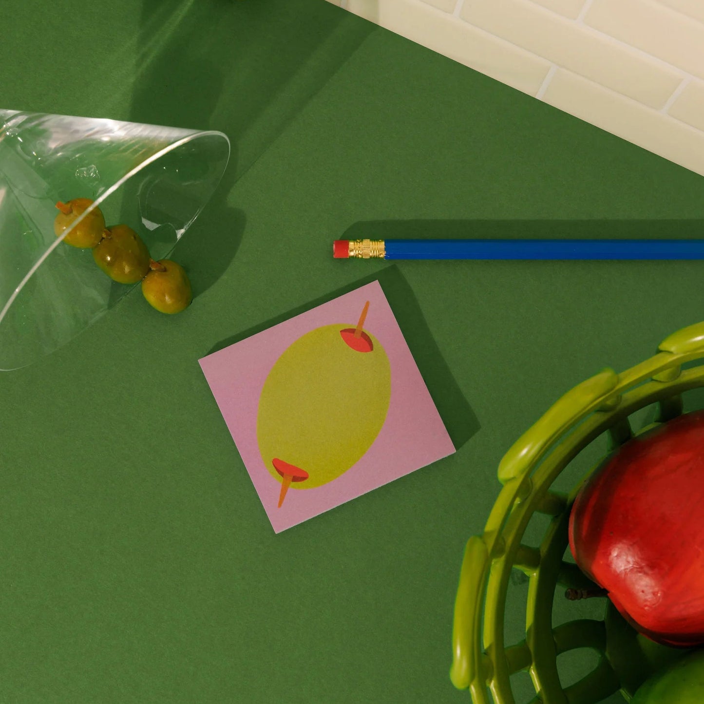 Colorful objects including a pencil, a small card with a green apple, and a basket with apples on a green surface.