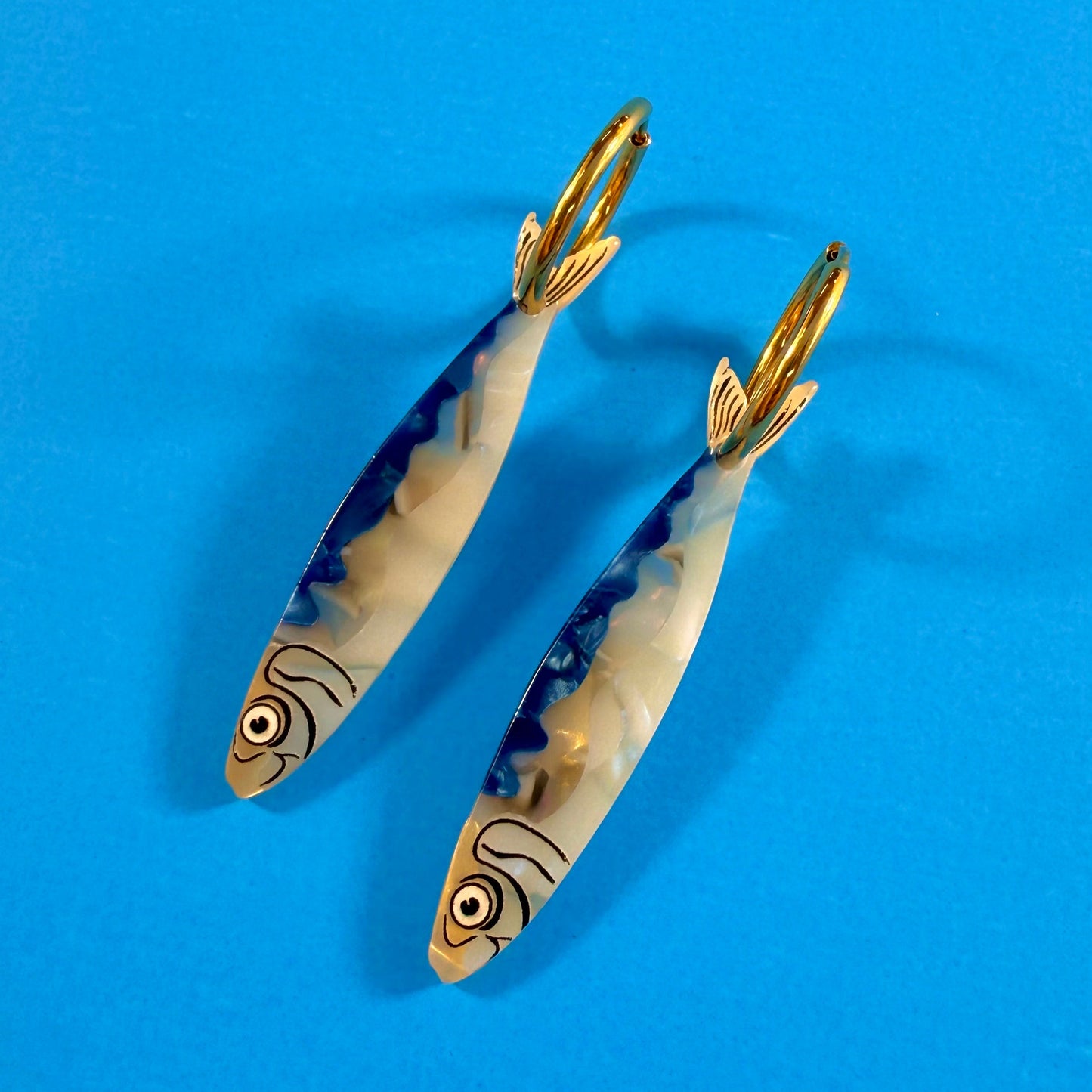 Sardine fish earring charms on gold hoops on top of a blue background.