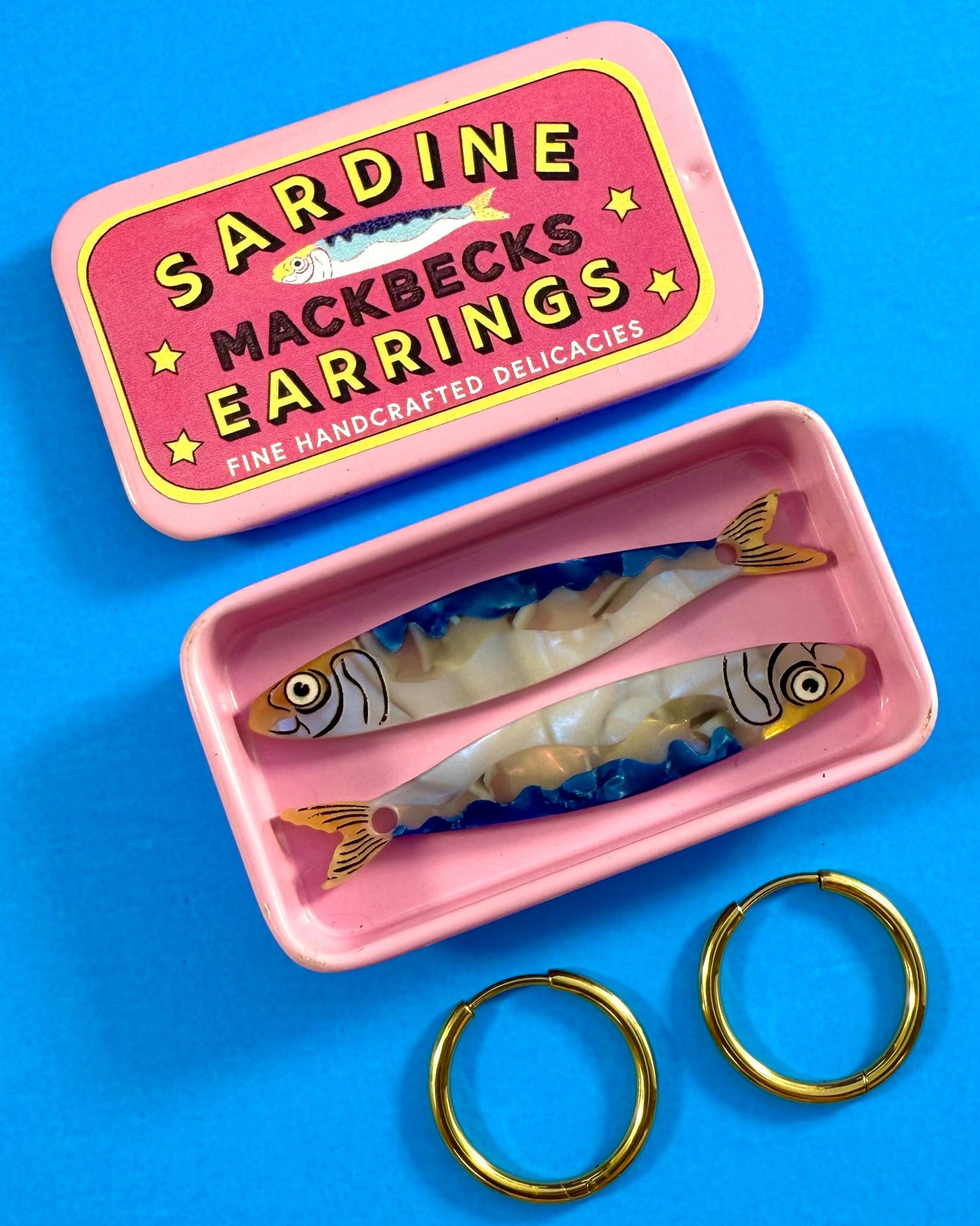 MackBecks Sardine Earrings packed in pink custom tin on a blue background.