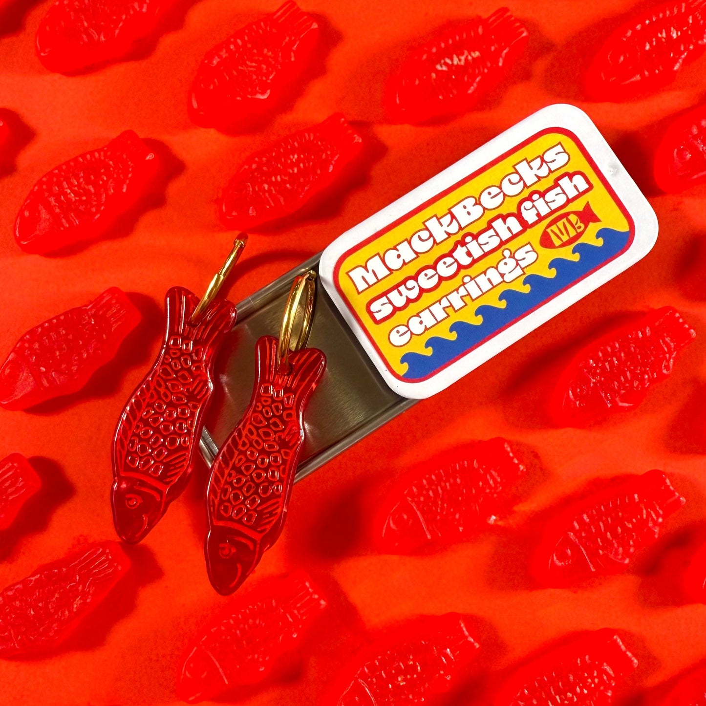 Red fish-shaped gummy candies with a MackBecks Sweetish Fish Earrings on tinned packaging on red background