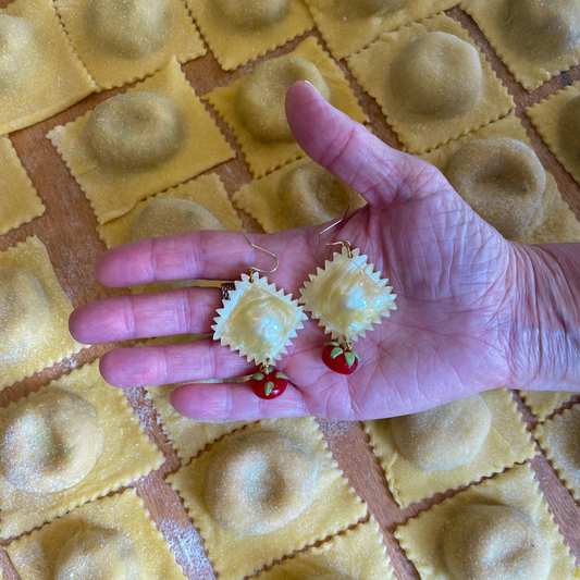 Hand holding a small ravioli earrings with glass tomato charms over a board of handmade pasta.