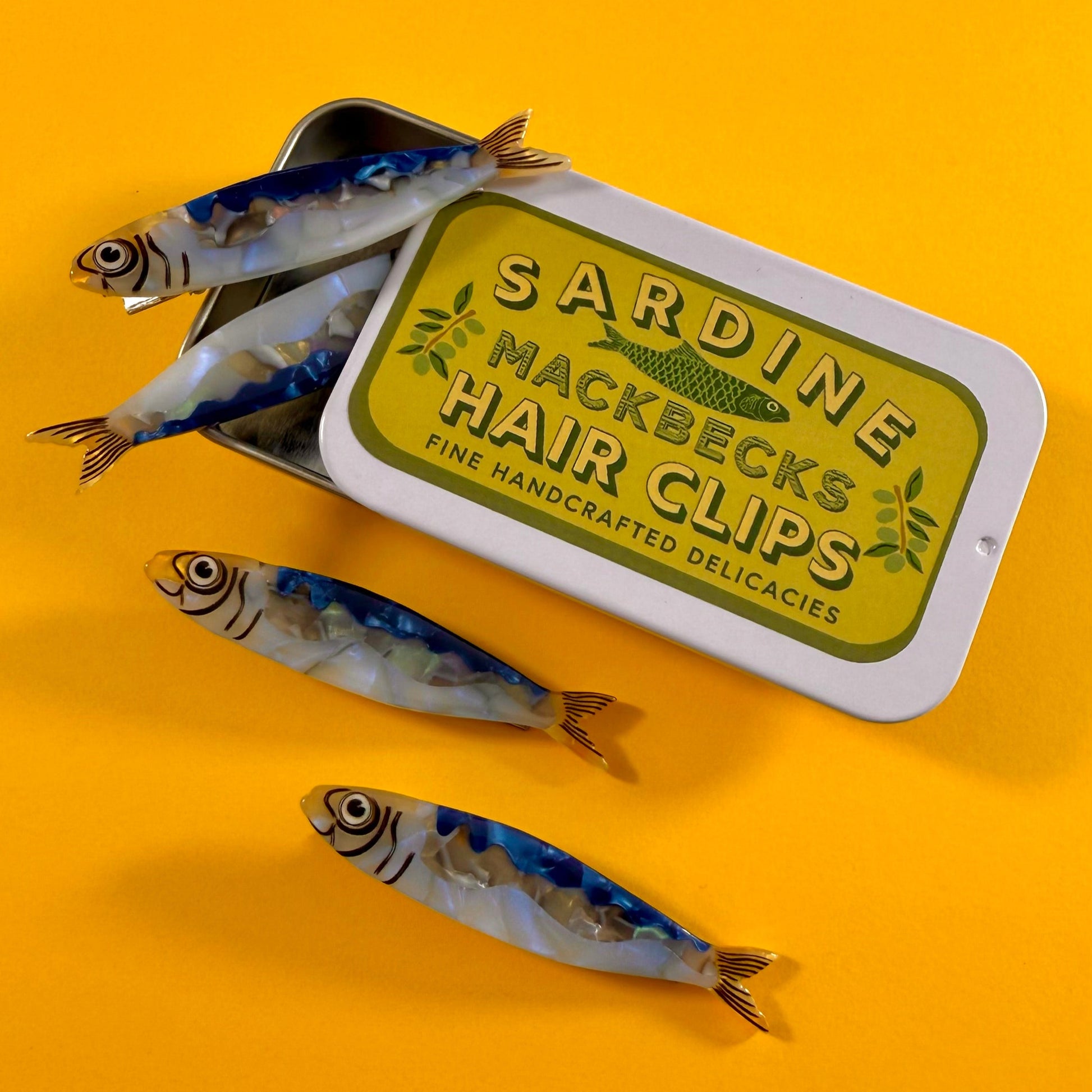Sardine-shaped hair clips with a tin labeled 'Sardine Mackbecks Hair Clips' on a yellow background