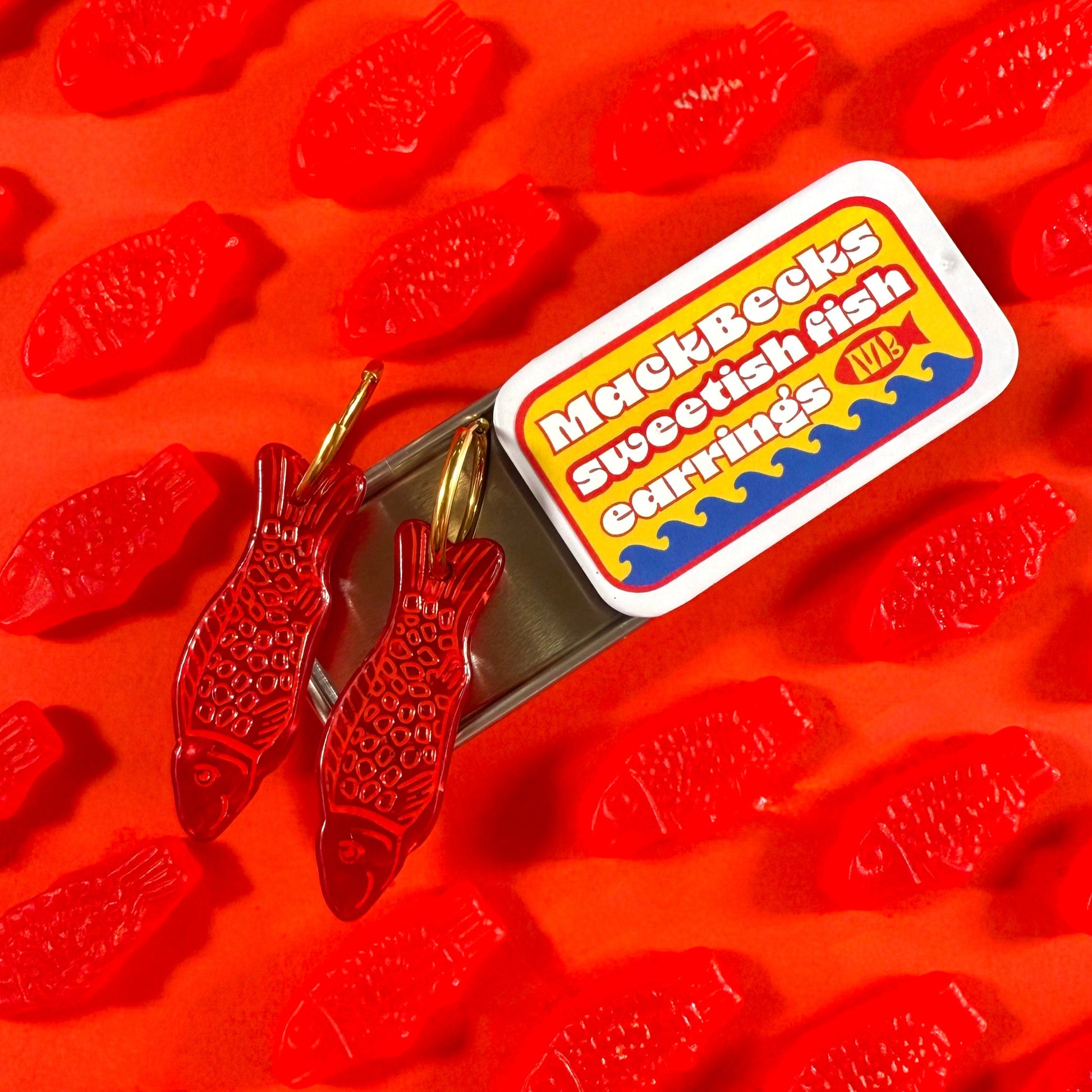 Red fish-shaped gummy candies with a MackBecks Sweetish Fish Earrings on tinned packaging on red background